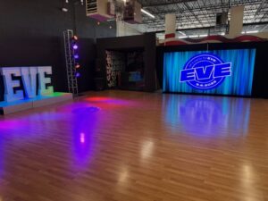 A spacious dance studio with wooden floors, colorful stage lights, and large illuminated letters spelling EVE. A blue-lit screen displays the EVE Academy logo beside the stage area.