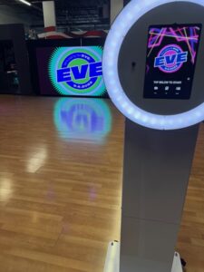 A photo booth with a glowing ring light stands on a wooden floor. In the background, a large circular logo that reads “EVE” is projected onto a screen, creating a colorful reflection on the floor.