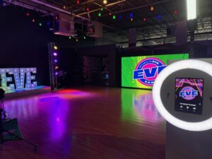 A spacious studio with wood flooring, colorful string lights overhead, a large EVE marquee, bright stage lights, and a digital display showing EVE branding on a green background. A photo booth is in the foreground.