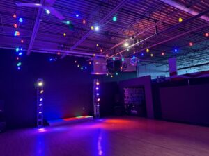 A spacious dance studio with a wooden floor, colorful string lights on the ceiling, stage lights, a small stage, a disco ball, and a dark wall with a poster in the background.
