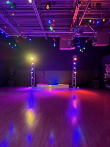 A spacious dance floor with colorful stage lights and string lights overhead, creating a vibrant atmosphere. A small stage or platform is at the back, and the wooden floor reflects the colorful lighting.