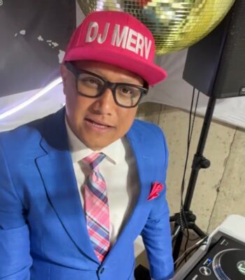 A man in a blue suit, pink plaid tie, and DJ MERV hat stands by DJ equipment, with a large disco ball in the background.